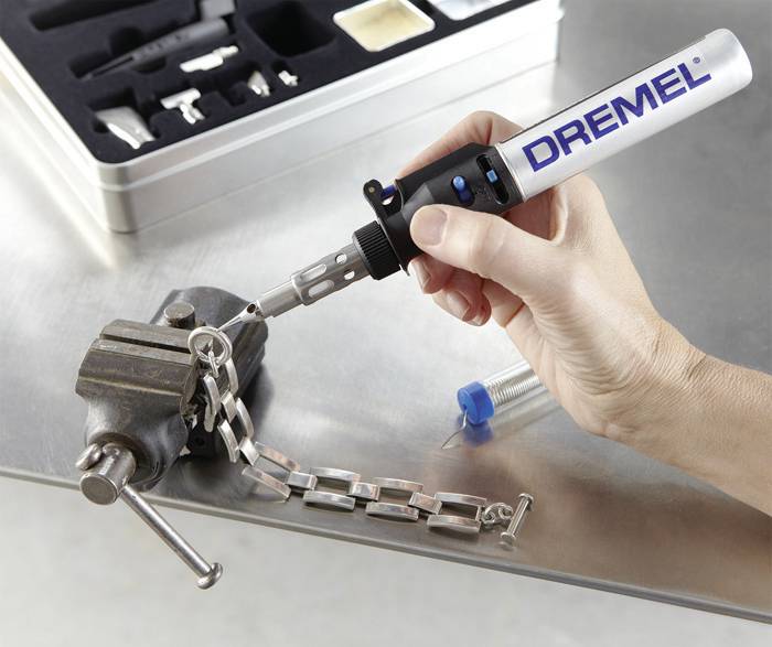 Eine Hand hält ein Dremel-Werkzeug und bearbeitet ein Metallarmband, das in einem Schraubstock auf einem Tisch fixiert ist.