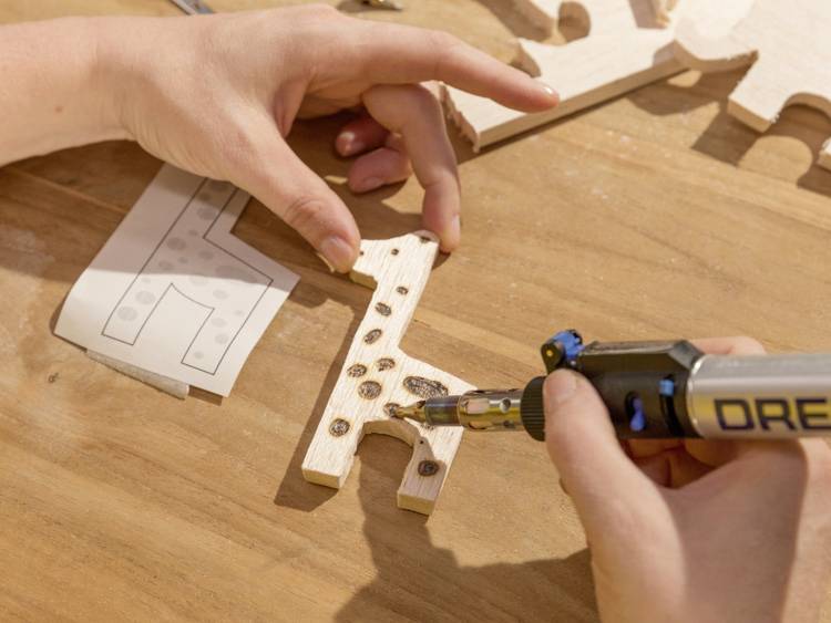 Person verwendet ein Lötkolben, um Löcher in ein hölzernes Puzzle-Stück zu brennen. Im Hintergrund sind weitere Puzzle-Teile.