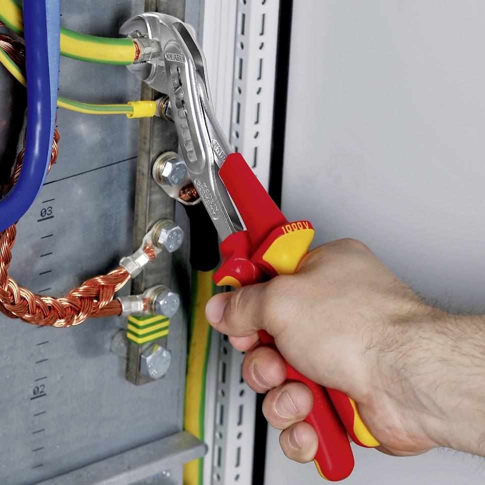 Eine Hand hält isolierte Zangen und einen Schraubenschlüssel, die an elektrischen Kabeln in einer Schalttafel arbeiten.