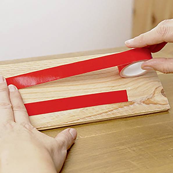 Eine Person bringt einen Streifen rotes Klebeband auf einer Holzoberfläche an und glättet ihn mit der Hand, was auf eine Bastel- oder Do-it-yourself-Aufgabe hindeutet.