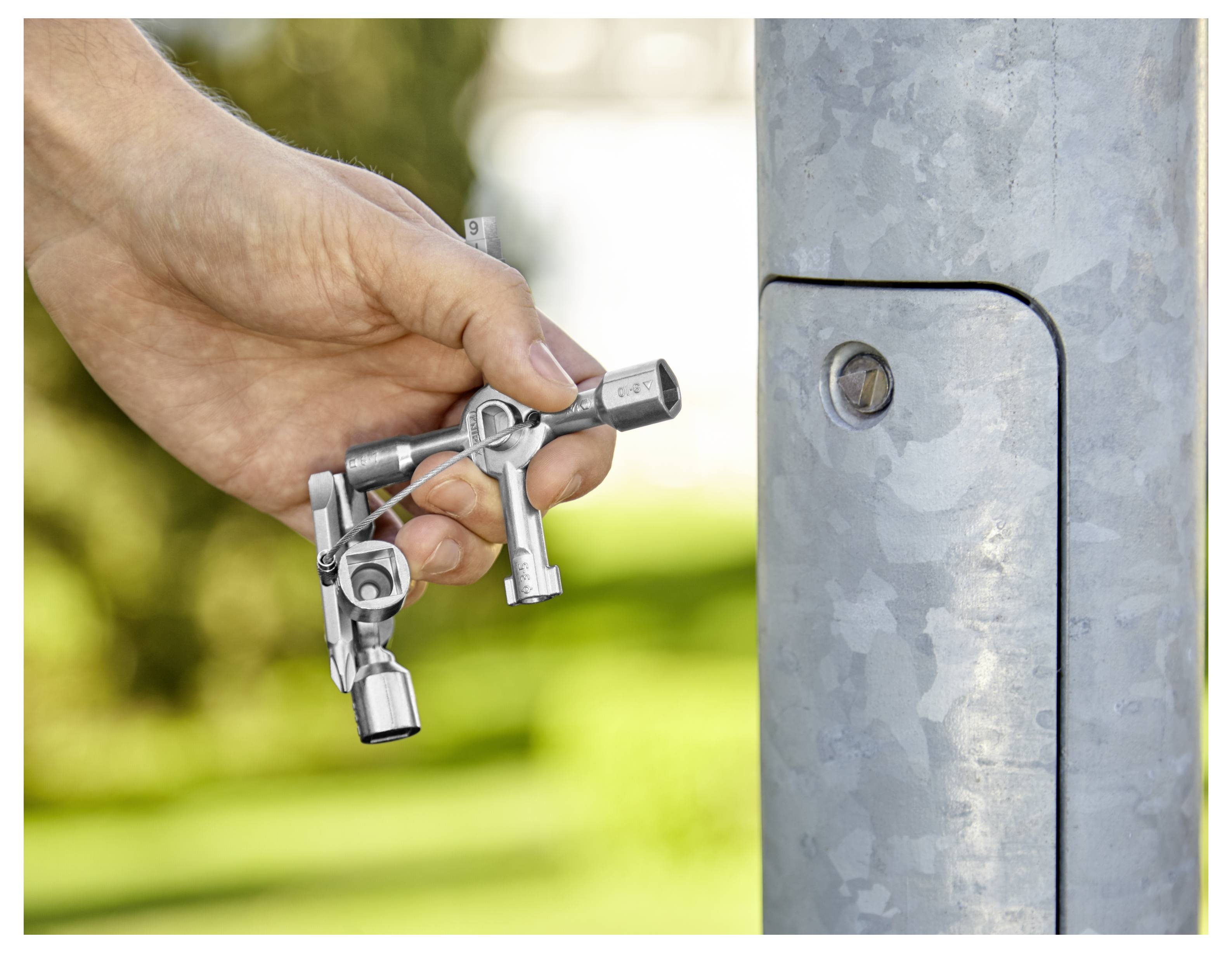 Eine Hand hält ein Metallwerkzeug in der Nähe einer Schraube an einem Metallpfosten, was auf Reparatur- oder Wartungsarbeiten hindeutet. Der Hintergrund ist im Freien und leicht unscharf.