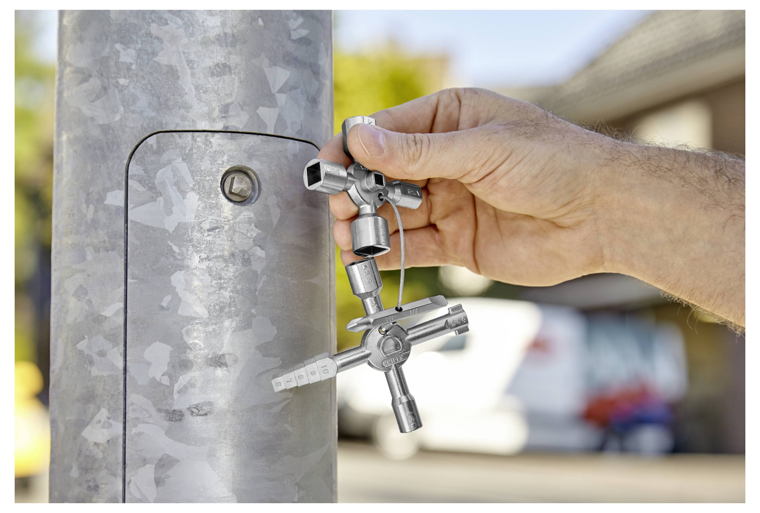 Eine Hand benutzt einen universalen Universalschlüssel, um ein verschlossenes Panel an einem Metallpfosten zu öffnen, draußen an einem sonnigen Tag mit unscharfem städtischem Hintergrund.