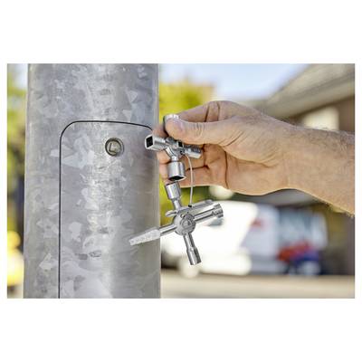 Eine Hand benutzt einen universalen Universalschlüssel, um ein verschlossenes Panel an einem Metallpfosten zu öffnen, draußen an einem sonnigen Tag mit unscharfem städtischem Hintergrund.