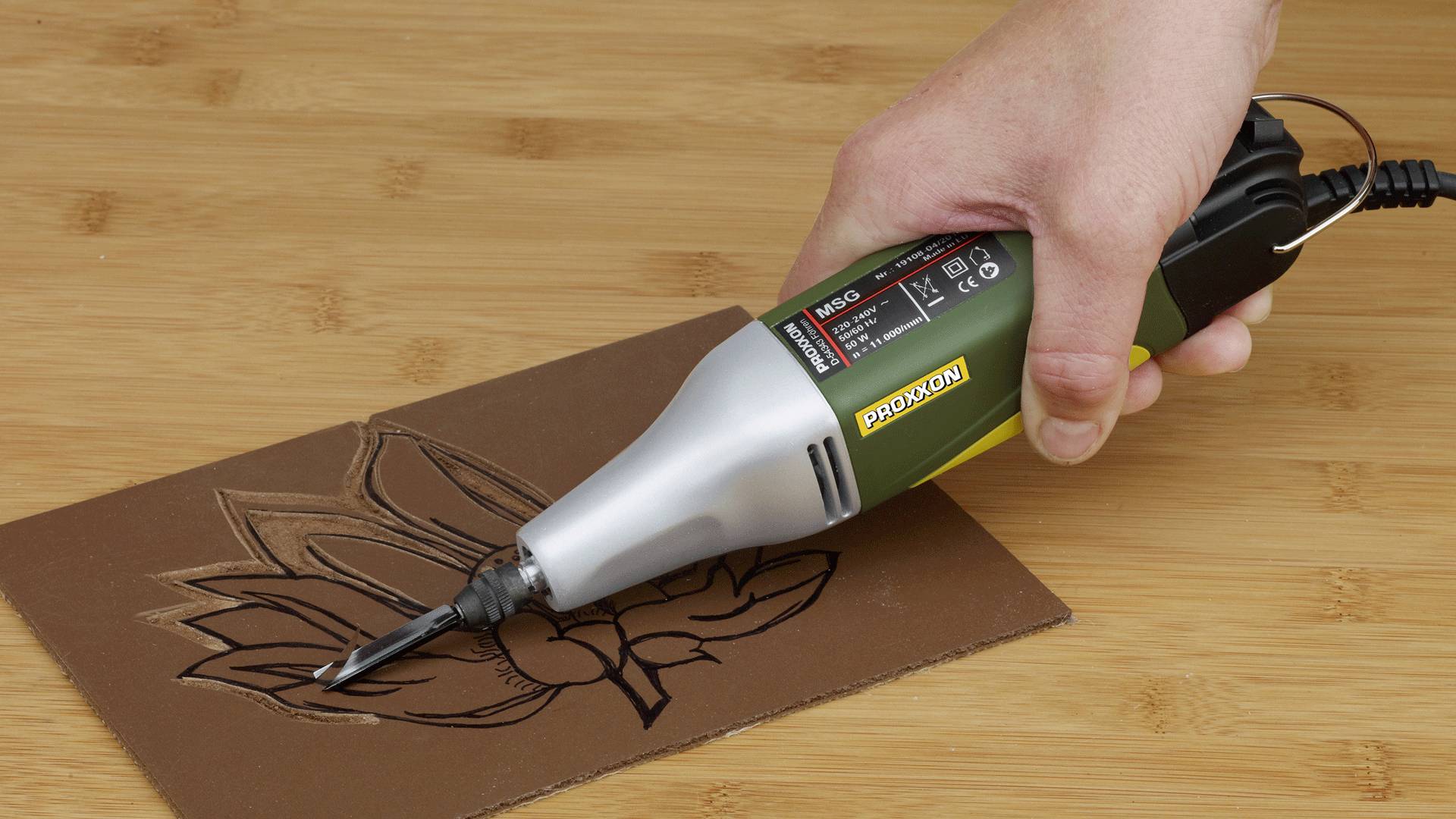 Eine Person verwendet einen elektrischen Graviergerät, um ein Blumendesign auf ein braunes Material zu gravieren, das auf einer Holzoberfläche liegt.
