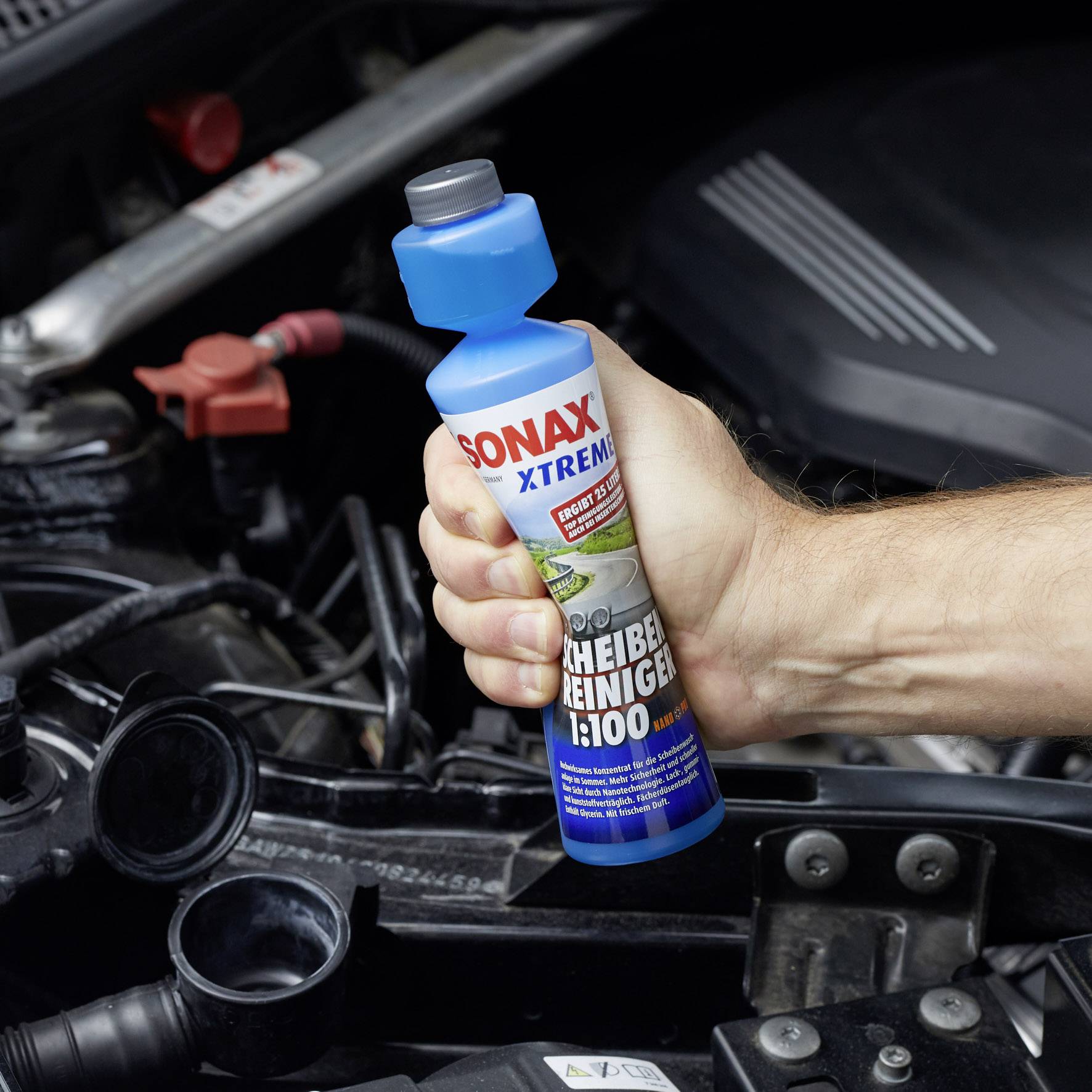 Eine Hand hält eine Flasche Scheibenreiniger neben einem Auto-Motorraum.