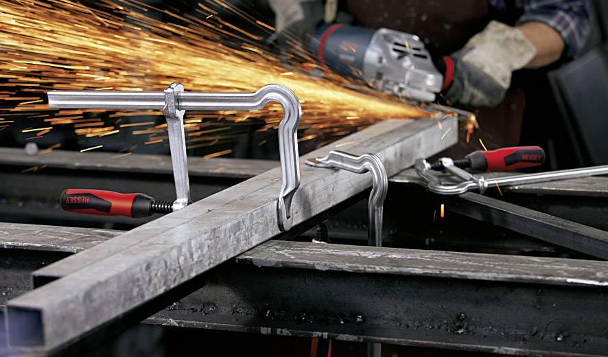 Eine Person arbeitet mit einem Winkelschleifer an einem Metallstück, Funken fliegen in die Luft. Klemmen halten das Metall stabil.