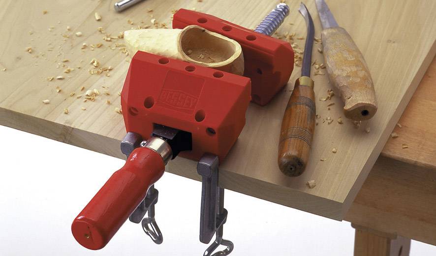 Rote Tischklemme hält Holzstück, umgeben von Schnitzwerkzeugen auf Holztisch. Holzspäne verstreut. Hauptmotiv: Holzbearbeitungswerkzeuge.