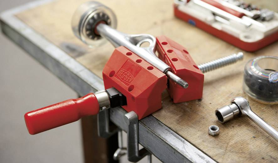 Eine Skateboard-Achse wird mit roten Schutzbacken in einem Schraubstock auf einem Werkbank befestigt. Im Hintergrund befinden sich Schrauben und Werkzeuge.