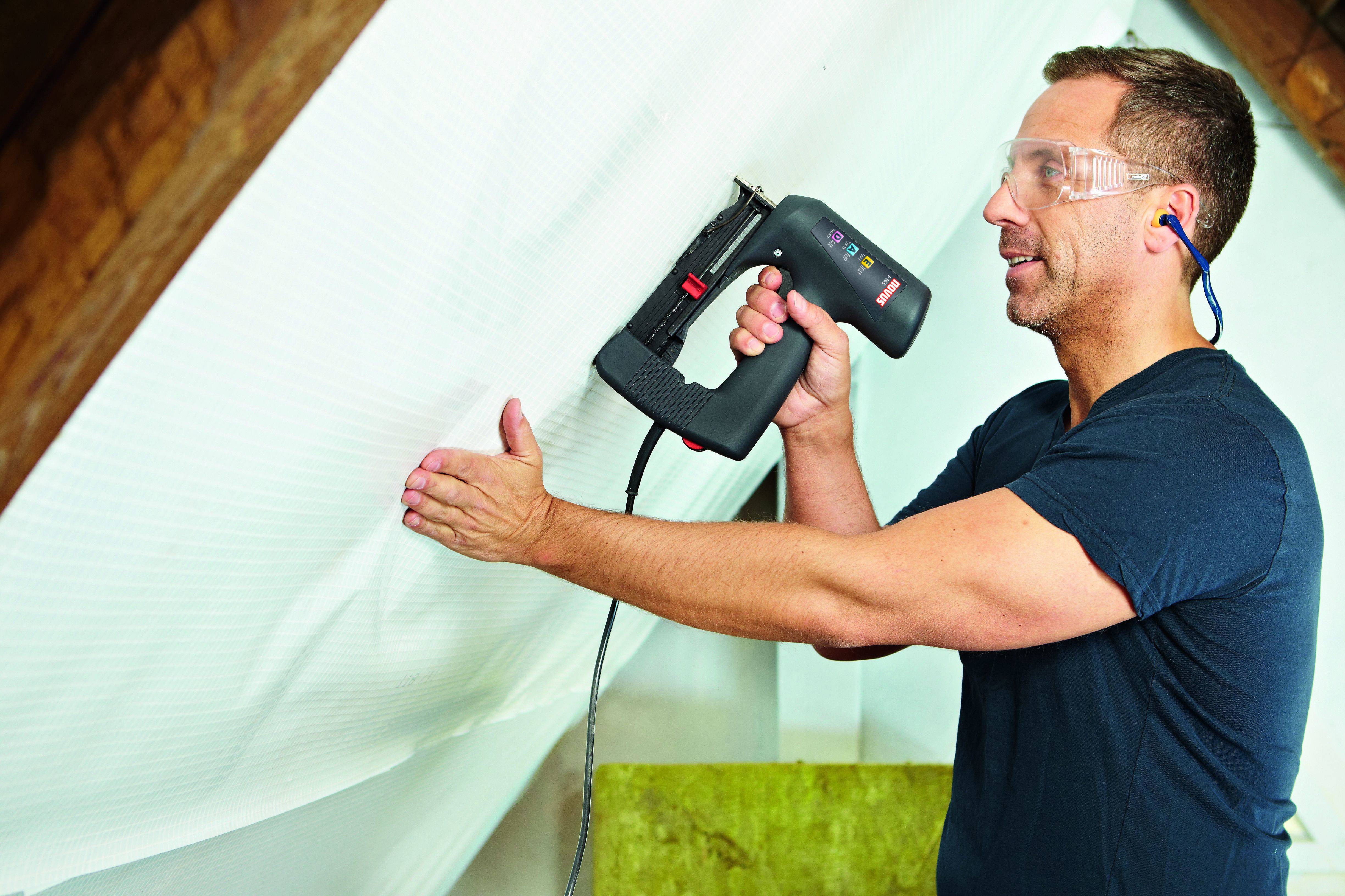 Ein Mann befestigt Dämmmaterial in einem Dachgeschoss, trägt Schutzbrille und Gehörschutz, hält einen elektrischen Tacker in der Hand.