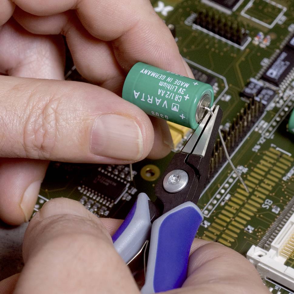 Eine Person setzt mit einer Zange eine grüne Batterie auf eine elektronische Platine ein und verbindet sie mit einem Draht.