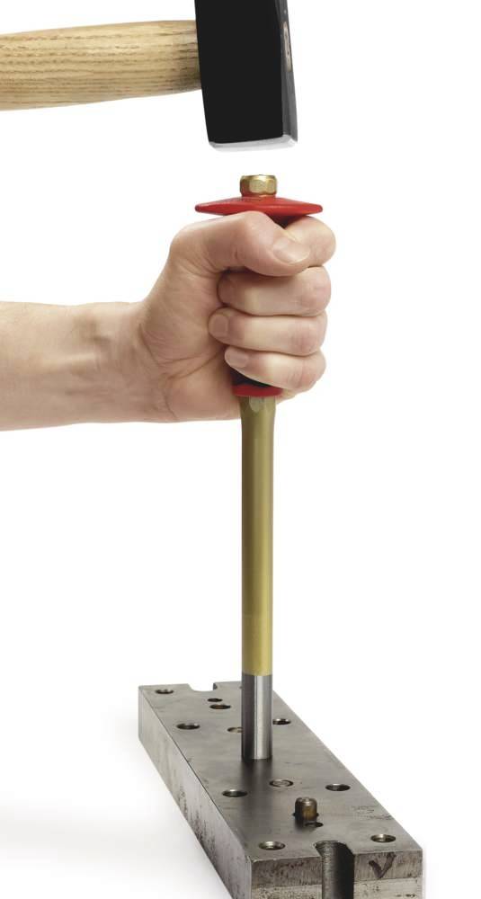 Eine Hand hält einen Meißel auf eine Metallplatte, während ein Hammer schlägt. Die Szene zeigt ein handwerkliches Bearbeitungsverfahren.