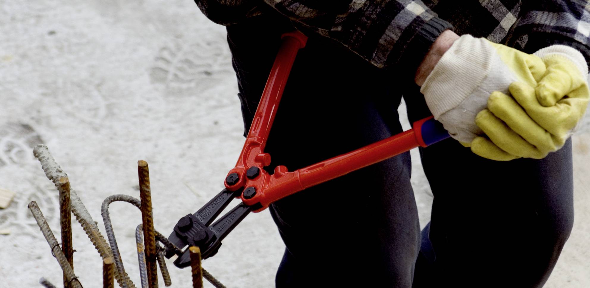 Eine Person mit gelben Handschuhen schneidet Metallstangen mit einem großen, roten Bolzenschneider auf einer Baustelle.