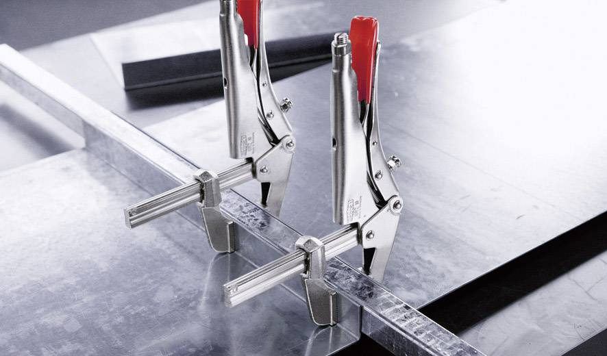 Zwei Klemmen halten Metallteile zusammen auf einem Arbeitstisch. Die Klemmen sind aus Metall mit roten Griffen, für präzise Arbeit.
