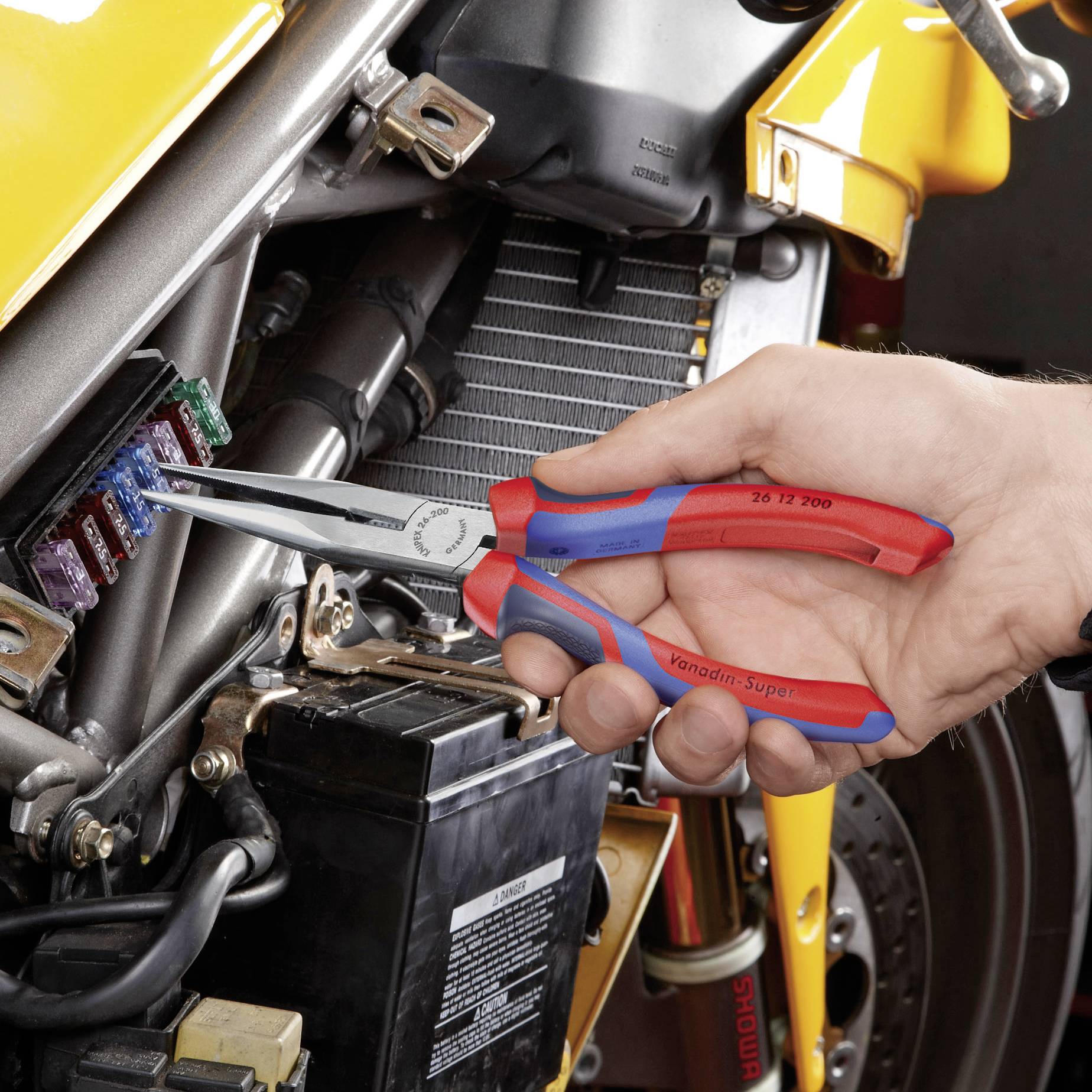 Eine Person hält eine rote und blaue Zange und entfernt eine Sicherung aus einem Sicherungskasten an einem gelben Motorrad.