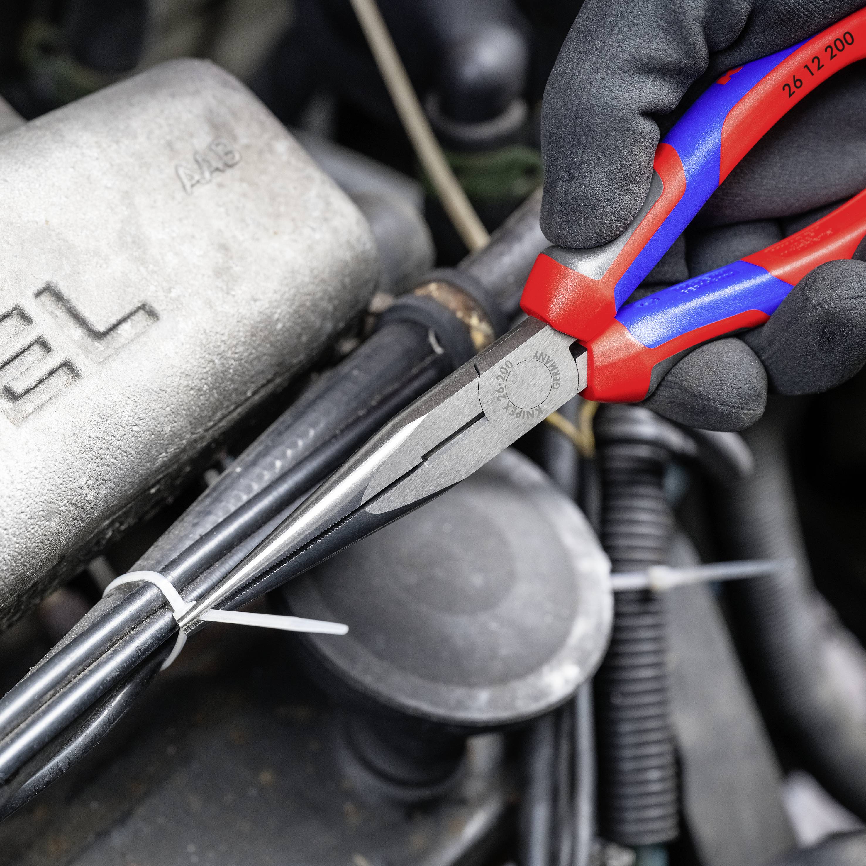 Ein Paar rot-blau isolierte Zangen schneidet einen Kabelbinder in einem Motorraum durch. Im Hintergrund ist ein Motorblock sichtbar.