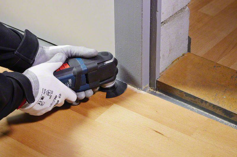 Eine Person verwendet ein Multifunktionswerkzeug, um einen präzisen Schnitt am Rand eines Holzbodens nahe einer Türöffnung durchzuführen.