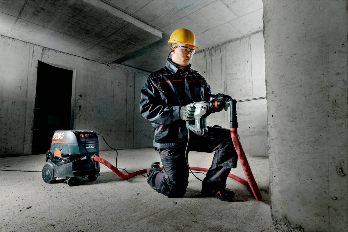 Eine Person in Schutzkleidung und Helm bohrt mit einem elektrischen Gerät in eine Betonwand in einem unfertigen Raum.