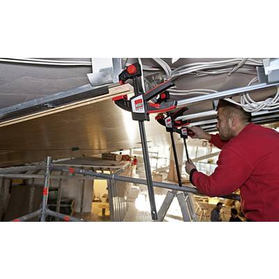 Ein Mann in roter Arbeitskleidung befestigt mit großen Klemmen eine Platte an einer Decke, umgeben von Kabeln und Baugerüsten.