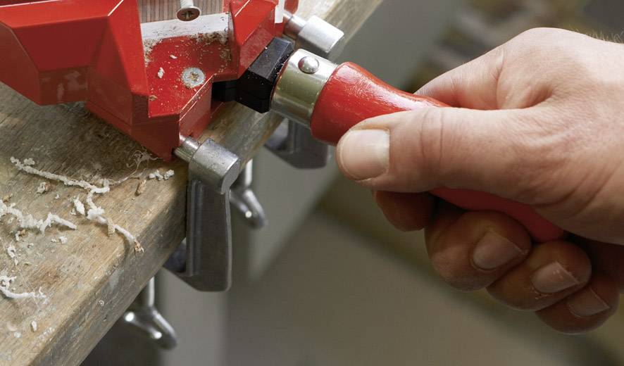 Eine Hand hält einen roten Griff, der ein Werkzeug anspannt, das an einem Holzstück fixiert ist. Holzspäne sind auf der Oberfläche zu sehen.