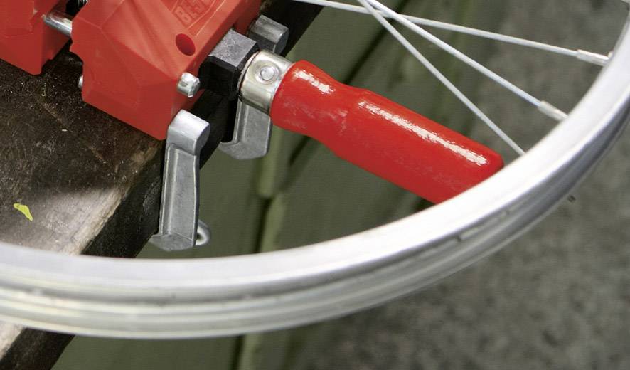 Ein silbernes Fahrradlaufrad wird mittels rotem Werkzeug bearbeitet, auf einer Werkbank fixiert.