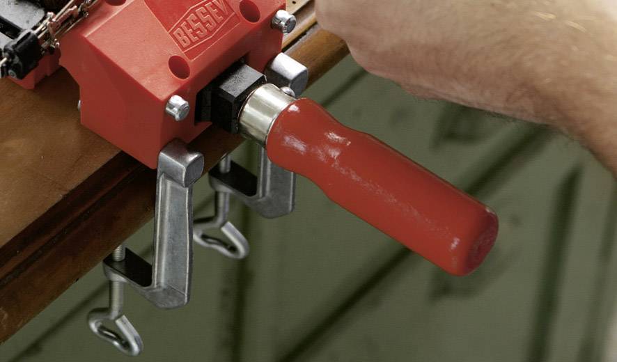 Eine Hand dreht einen roten Griff einer Schraubzwinge, die an einem Holztisch befestigt ist.