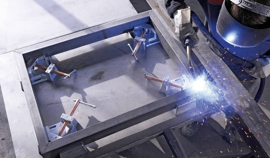 Eine Person schweißt ein Metallrahmenstück mit Funkenflug. Das Werkstück ist in einer Werkstatt montiert und mit Halterungen gesichert.