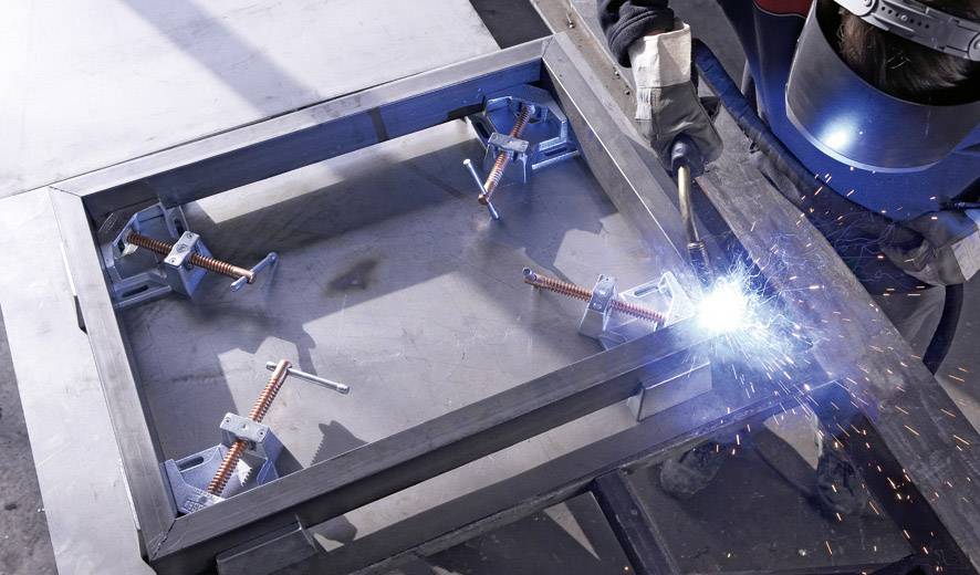 Ein Schweißer mit Schutzhelm arbeitet an einem Metallrahmen und erzeugt Funken. Der Rahmen ist auf einer Werkbank fixiert.