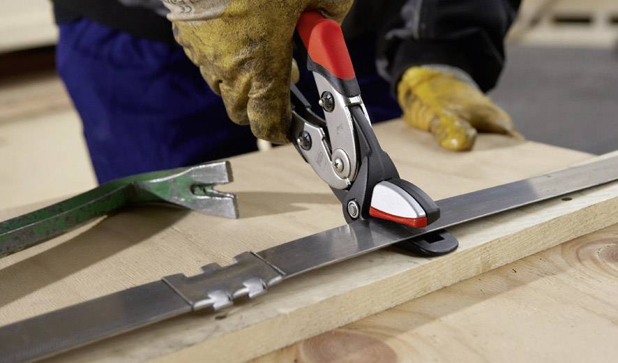 Person schneidet mit einer Metallschere ein Stahlband auf einem Holzbrett. Trägt gelbe Handschuhe für Schutzmaßnahmen.