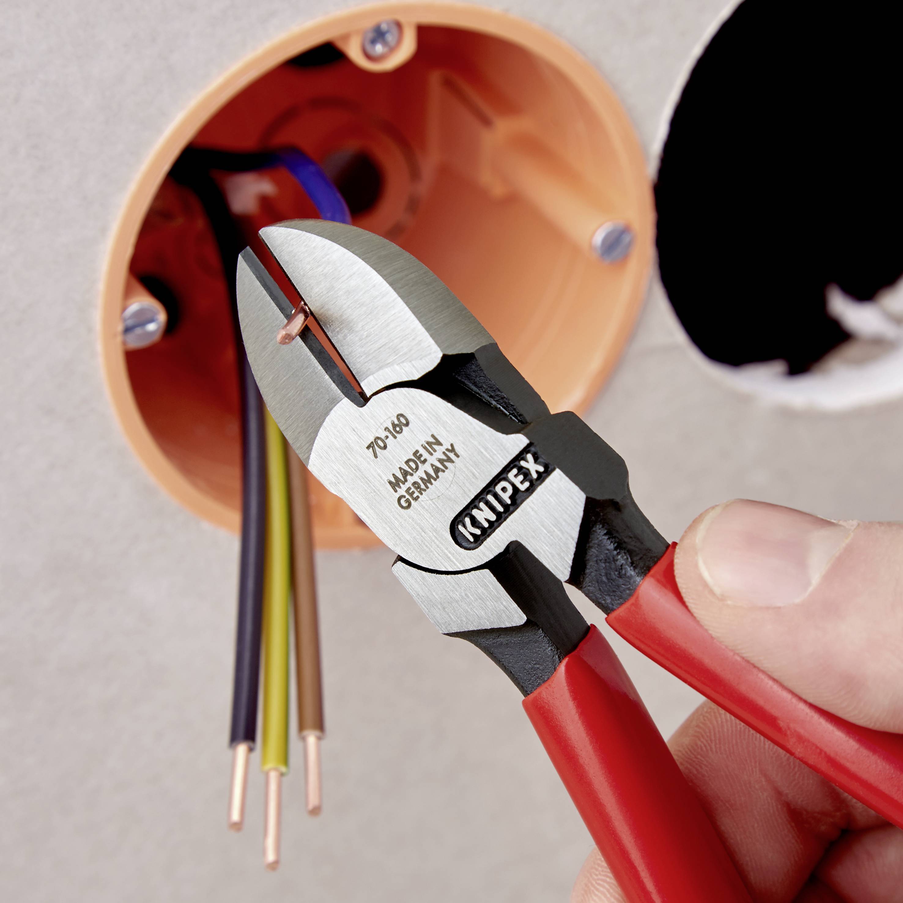 Eine Hand hält eine rote Zange und schneidet Drähte in einer offenen Steckdose. Die Zange zeigt 'Knipex Germany'.