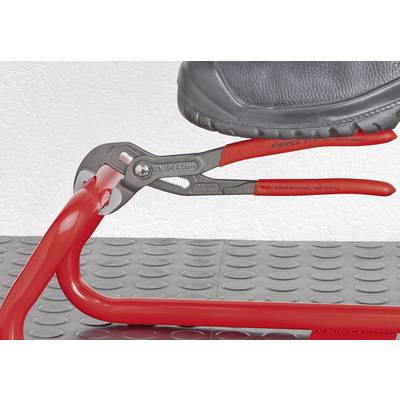 Ein Schuh steht auf einer roten Zange, die ein rohrförmiges Metallteil umfasst, auf einer gepunkteten Bodenplatte.