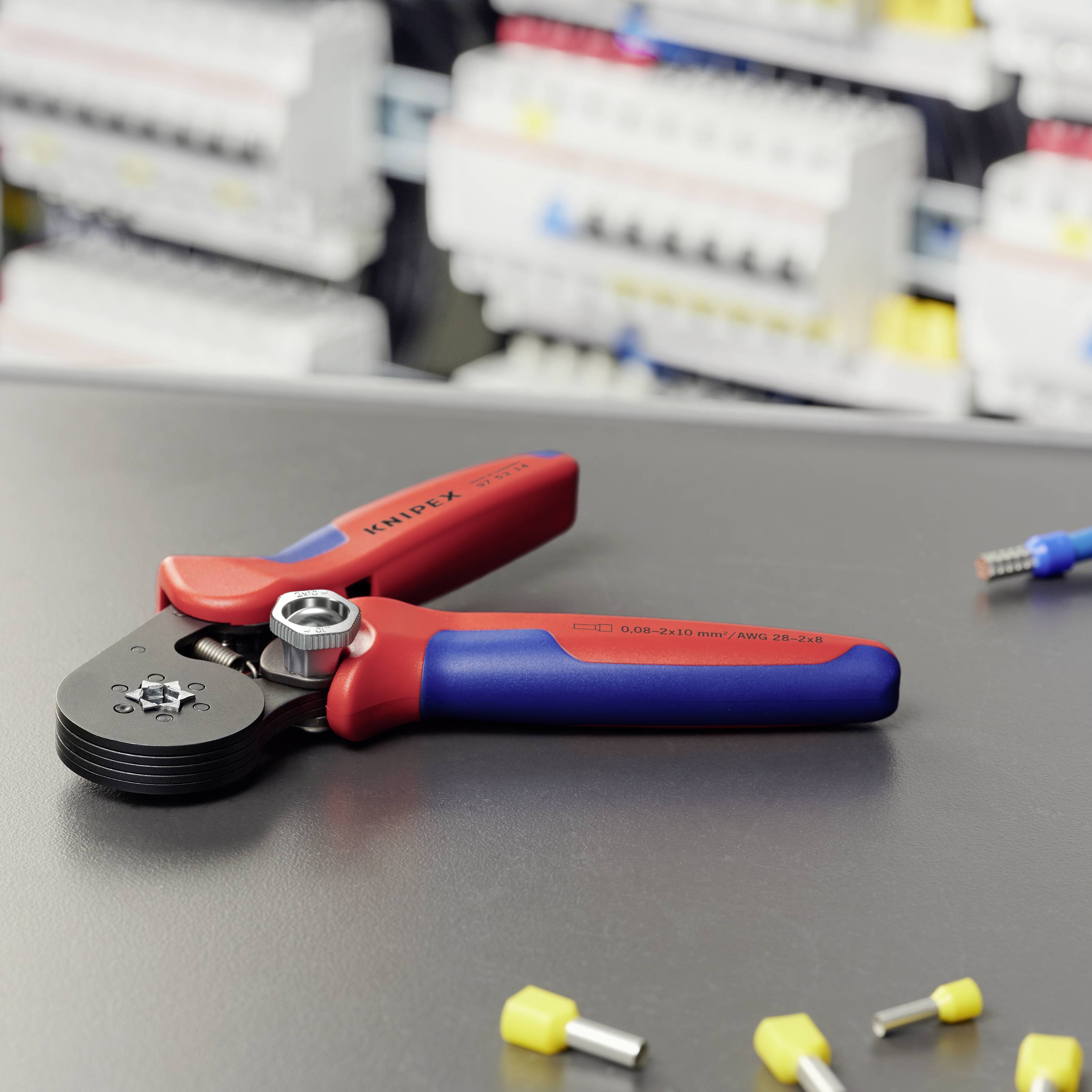 Eine rote und blaue Crimpzange liegt auf einem Tisch, umgeben von Kabelschuhen. Im Hintergrund ist eine verschwommene Schalttafel zu sehen.