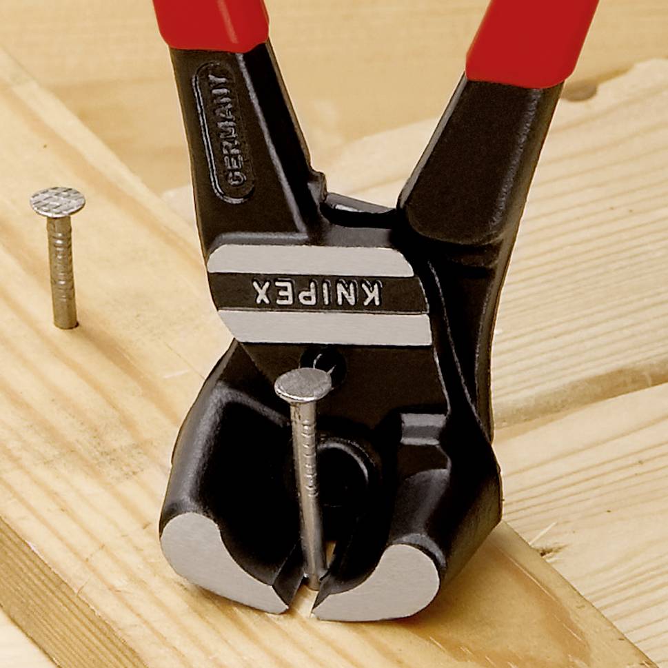 Eine Beißzange wird verwendet, um einen Nagel aus einem Holzbrett zu ziehen. Im Hintergrund sind zwei weitere Nägel zu sehen.