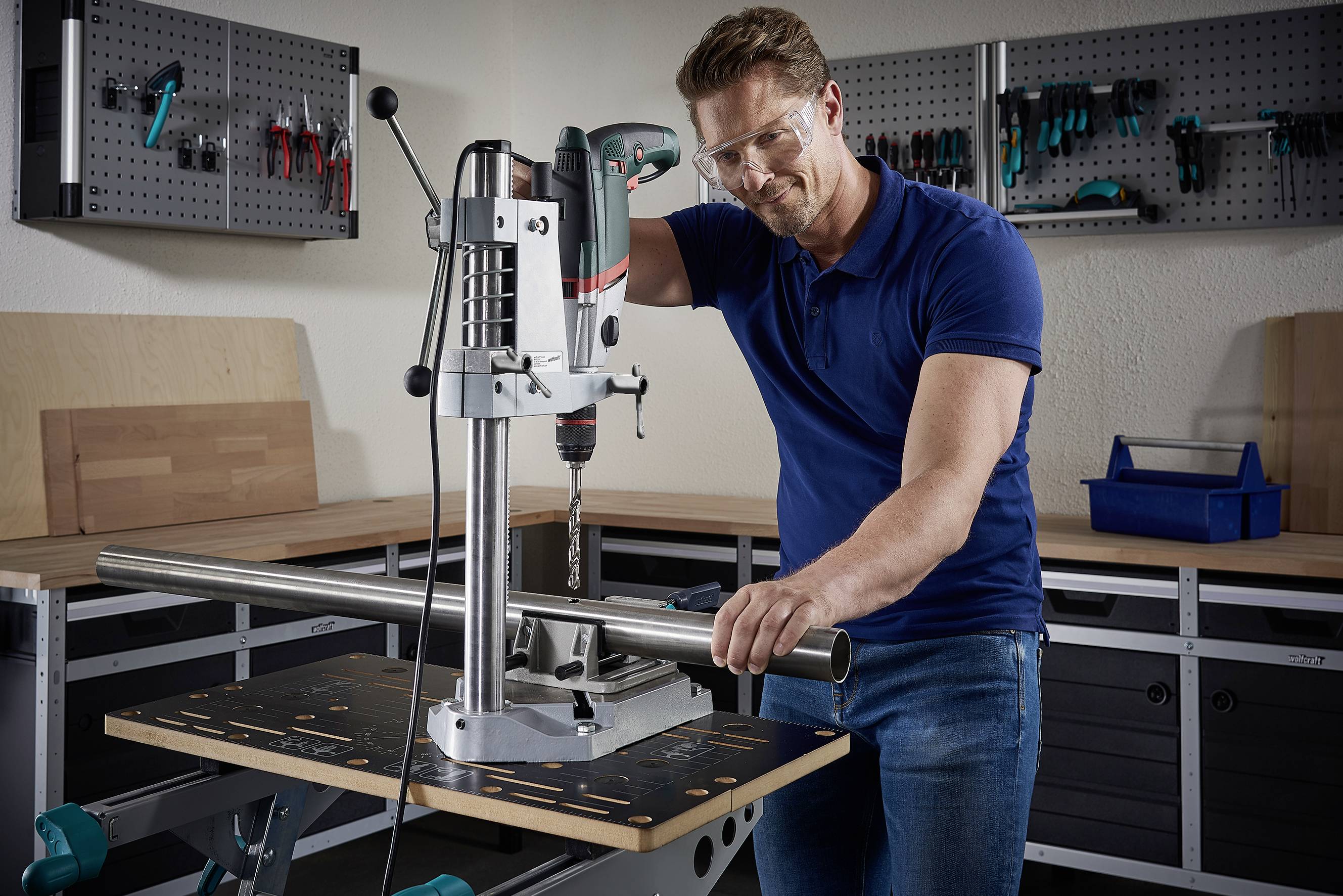 Ein Mann in Schutzbrille arbeitet an einer Tischbohrmaschine in einer Werkstatt und bearbeitet ein Metallrohr.