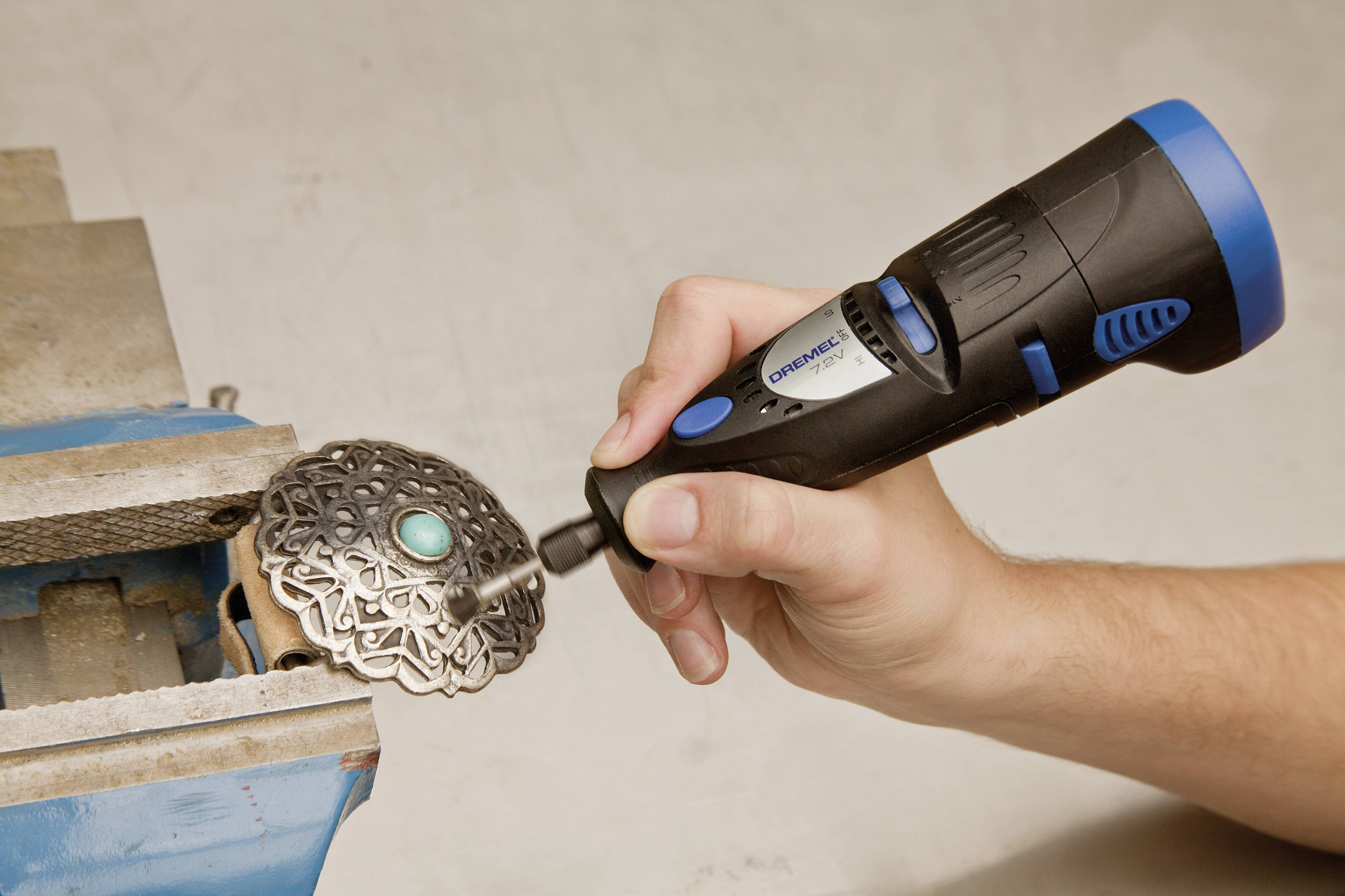 Eine Hand hält ein rotierendes Werkzeug und bearbeitet ein dekoratives Metallstück mit einem grünen Stein, das in einem Schraubstock eingespannt ist.
