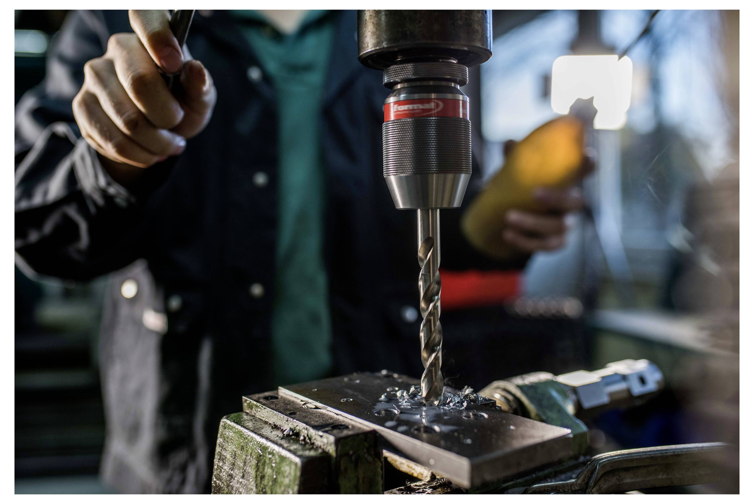 Nahaufnahme einer Person, die eine Bohrmaschine bedient und dabei ein in einem Schraubstock eingespanntes Metallwerkstück bearbeitet, wobei Metallspäne sichtbar sind.