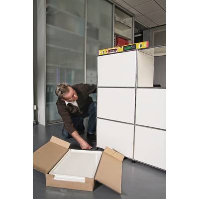 Ein Mann stellt einen Schrank mit Hilfe einer Wasserwaage und einer Wasserflasche ein. Ein geöffneter Karton liegt davor.