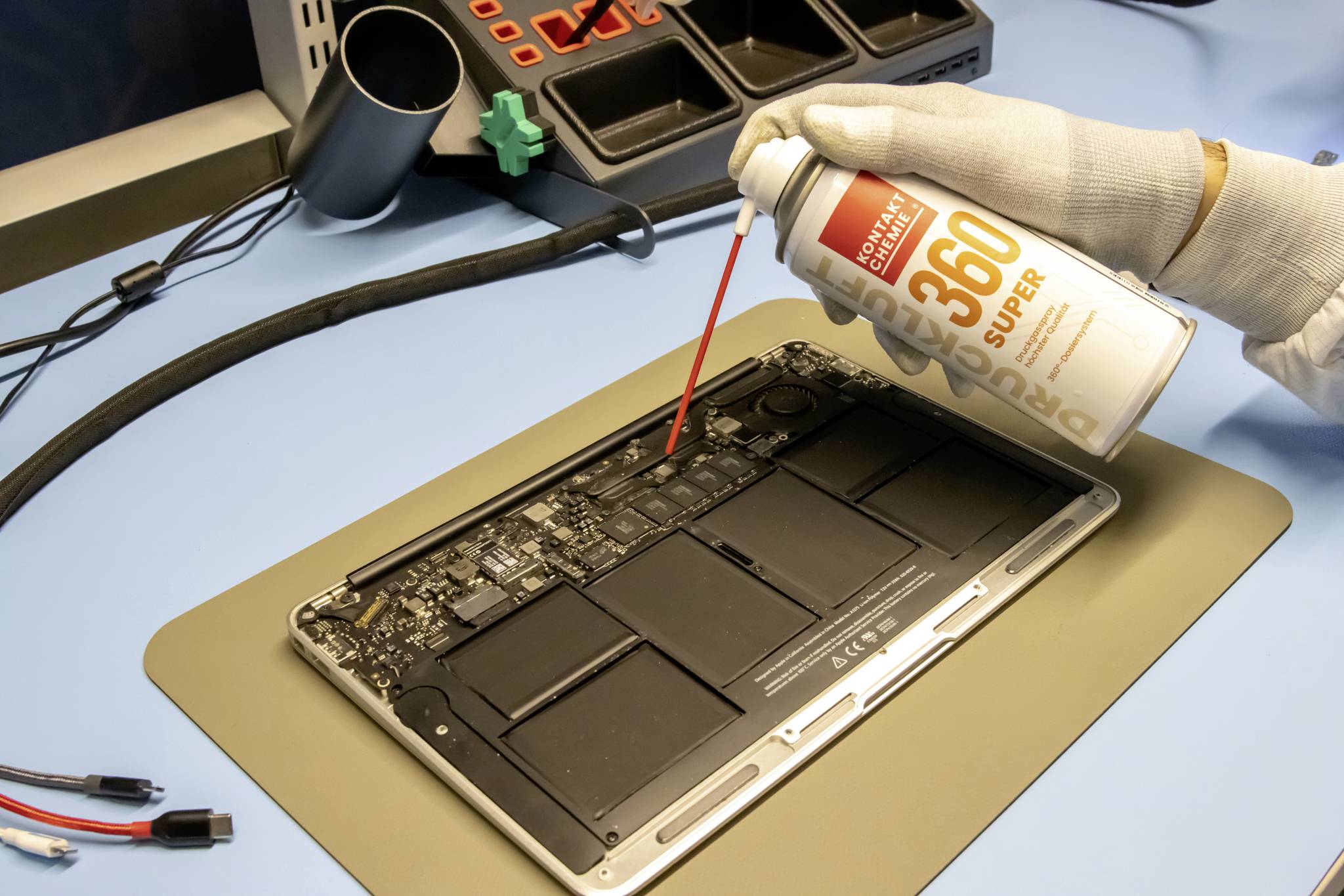 Person in Handschuhen reinigt die Innenseite eines geöffneten Laptops mit Druckluftspray auf einem Tisch mit Werkzeugen.