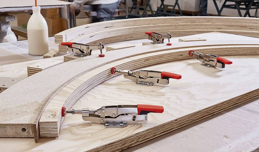 Holzwerkstatt mit gebogenen Sperrholzteilen, die von mehreren Klemmen auf einer Werkbank fixiert werden. Hintergrund unscharf.