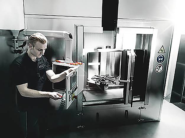 Eine Person bedient eine große Industriemaschine in einer Fabrikhalle und justiert Steuerungen an einem Kontrollpanel. Sicherheitshinweisschilder sind an der Maschine deutlich sichtbar.