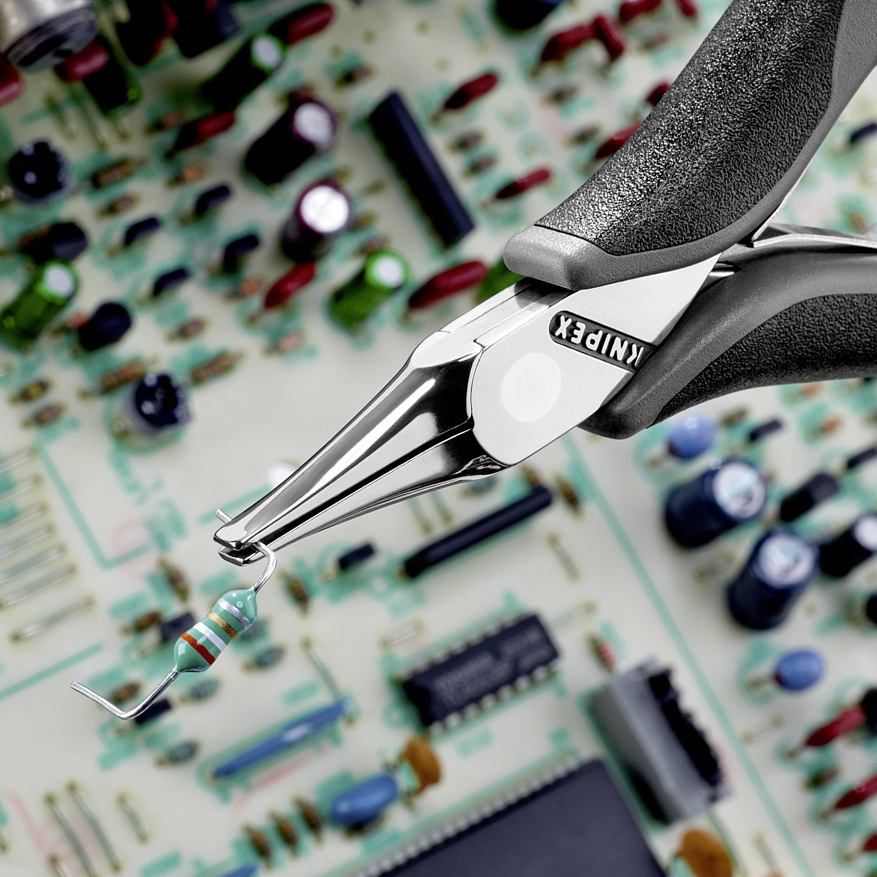 Eine Zange hält einen Widerstand vor einer elektrischen Leiterplatte mit verschiedenen Bauteilen.