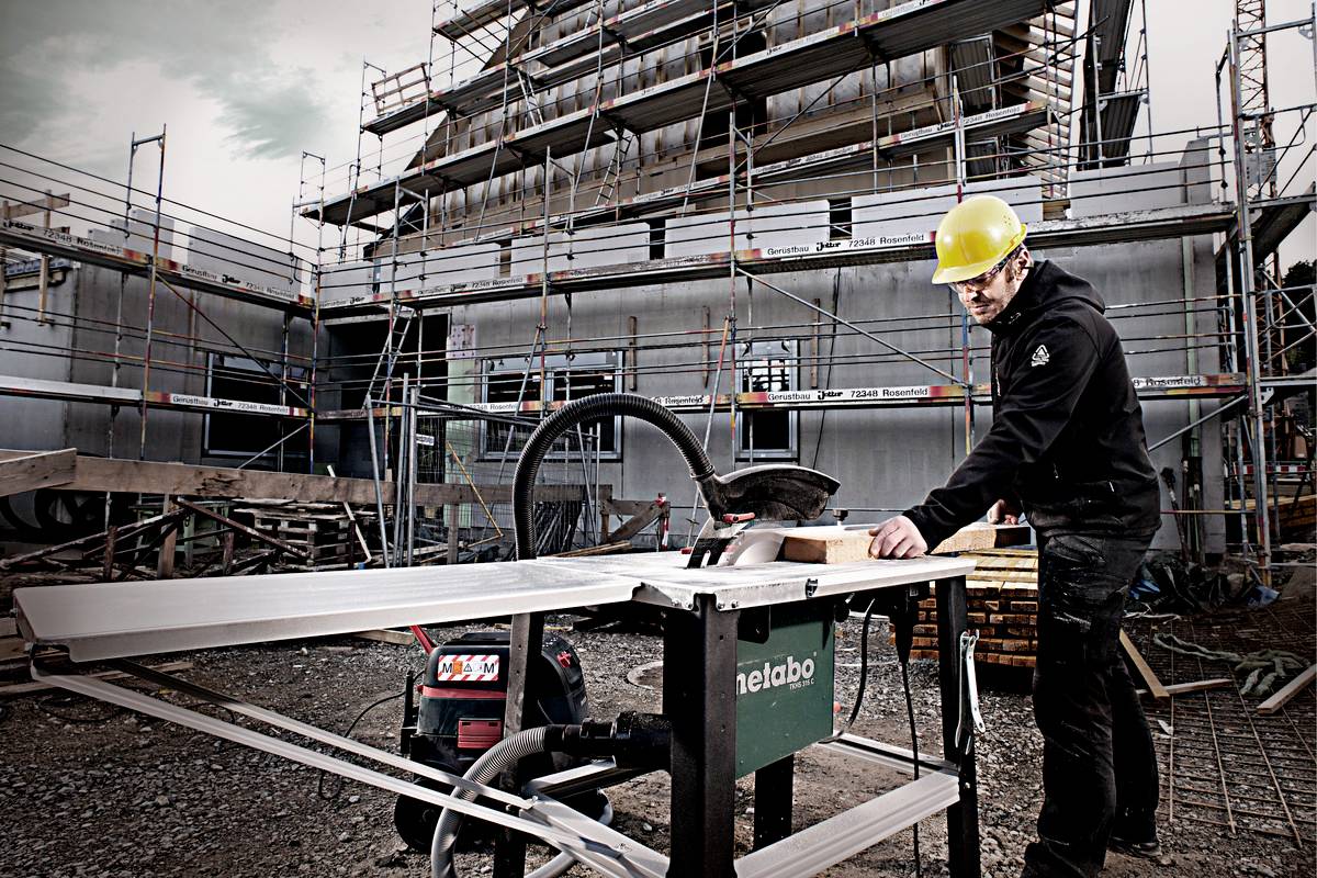 Ein Bauarbeiter mit gelbem Helm bedient eine Tischkreissäge auf einer Baustelle vor einem im Bau befindlichen Gebäude.