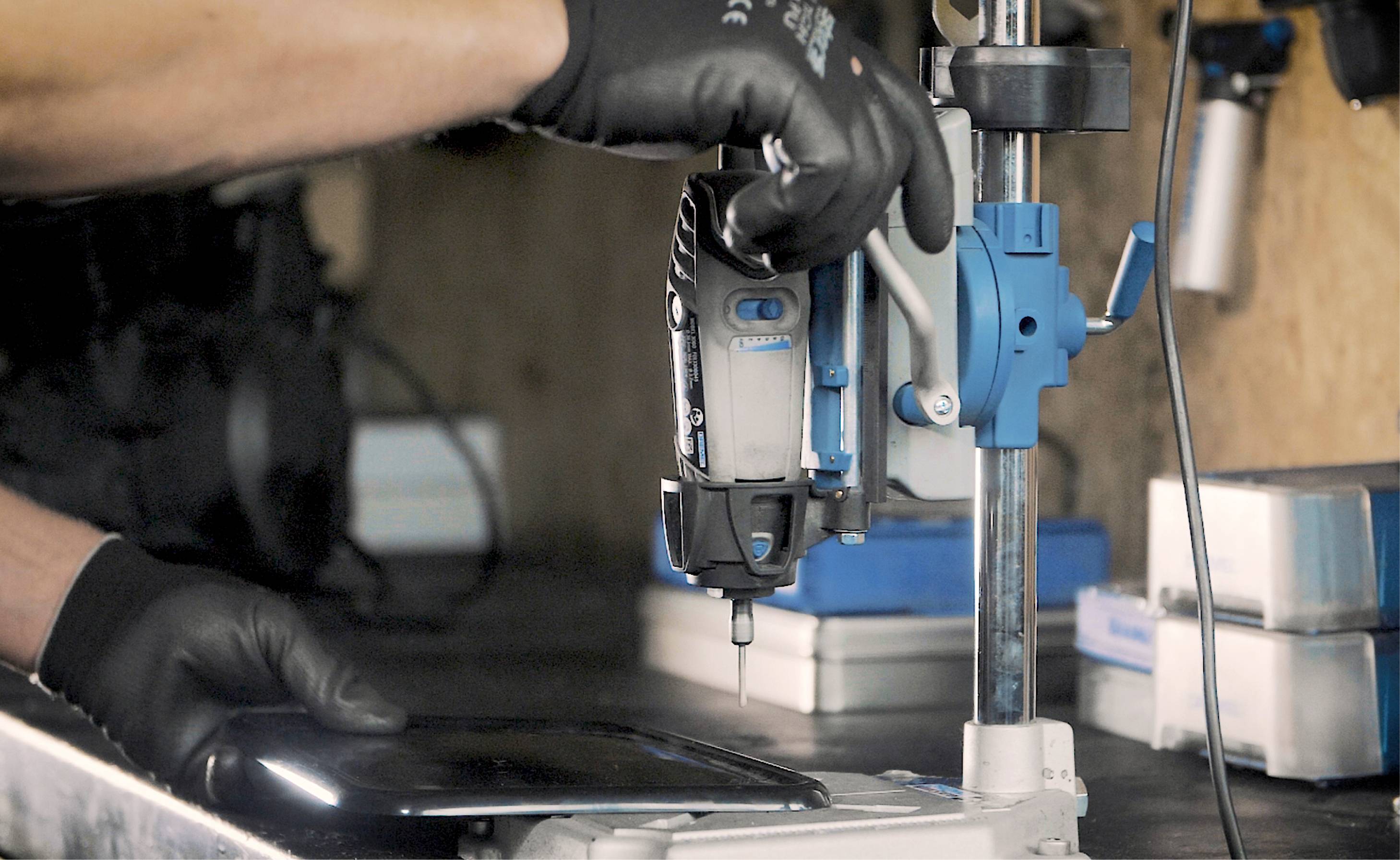 Eine Person mit Handschuhen bedient eine Bohrmaschine auf einem Tisch in einer Werkstattumgebung.