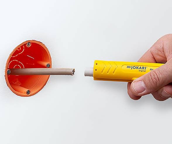 Eine Hand verwendet ein gelbes Abisolierwerkzeug, um die Isolierung von einem Kabel zu entfernen, das aus einer orangefarbenen Wandsteckdosenöffnung herausragt.