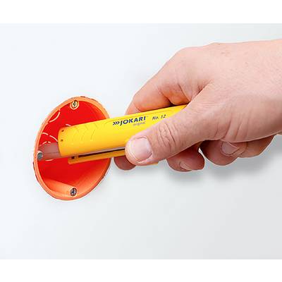 Eine Hand hält ein gelbes Kabelabisolierwerkzeug mit der Aufschrift "Jokari" und bereitet sich darauf vor, einen orangefarbenen Draht in einer Wand abzuisolieren.