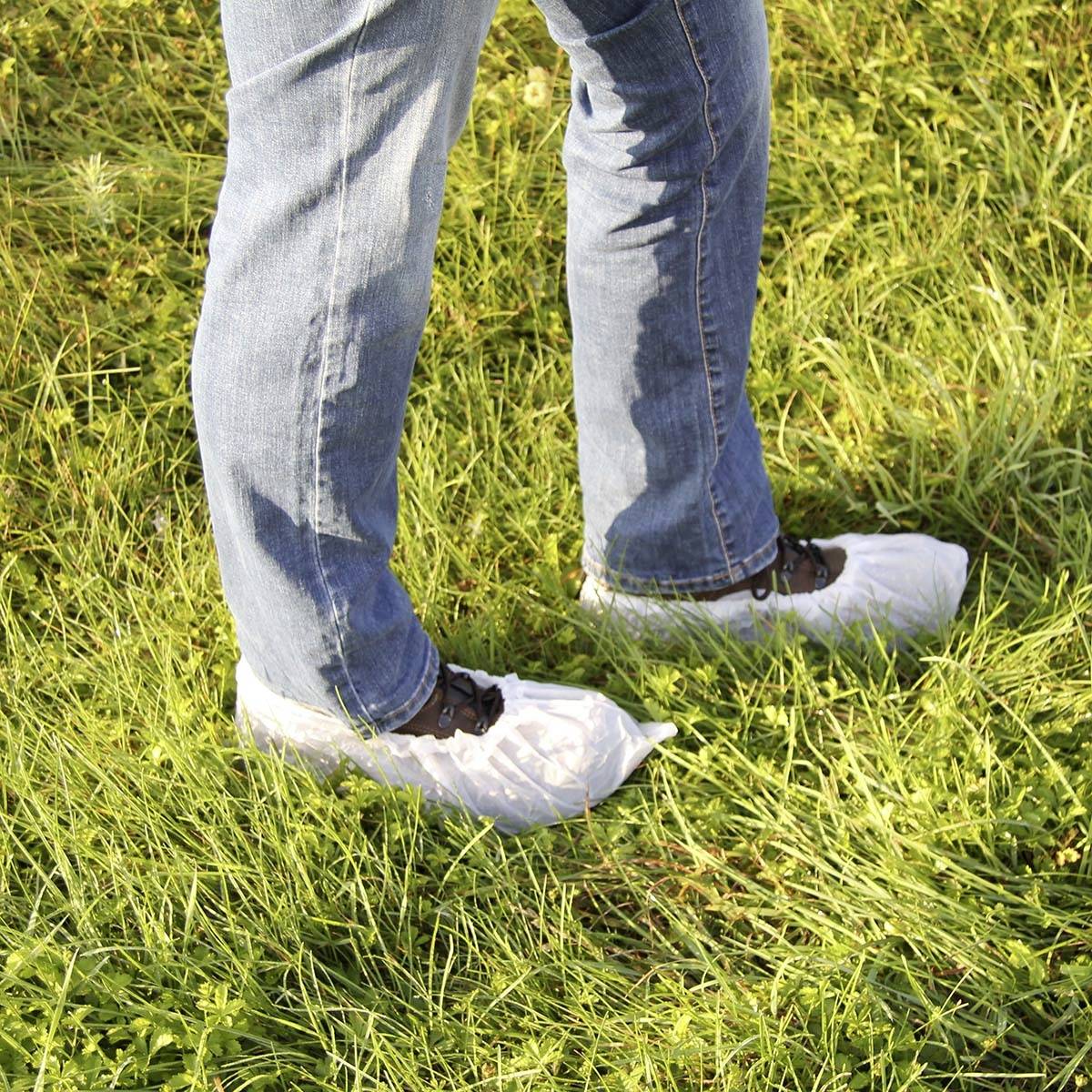 Ein Paar Beine in blauen Jeans stehen auf einer Wiese. Die Schuhe sind mit weißen Schutzüberzügen bedeckt.