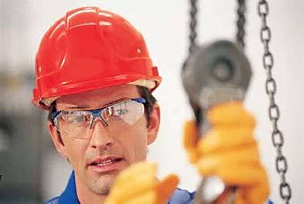 Ein Bauarbeiter mit rotem Schutzhelm und Schutzbrille steht vor unscharfem Hintergrund und bedient eine Kette mit einem Hebezeug.