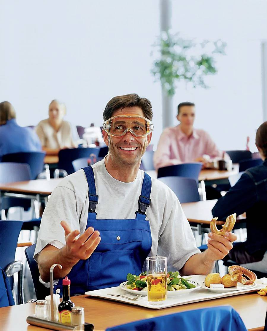 Arbeiter in blauem Overall und Schutzbrille sitzt lächelnd in der Kantine, isst einen Salat und hält ein Brötchen.