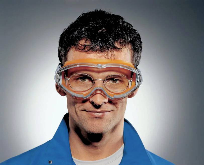 Eine Person trägt eine Schutzbrille und einen blauen Kittel. Hintergrund ist neutral, die Person lächelt leicht.