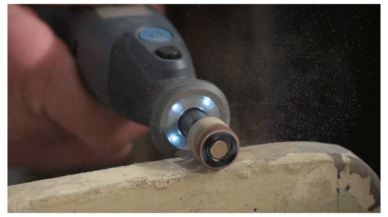 Eine Nahaufnahme eines Elektroschleifers, der eine Oberfläche bearbeitet, mit sichtbaren Staubpartikeln in der Luft, die den Glättungsprozess unterstreichen.