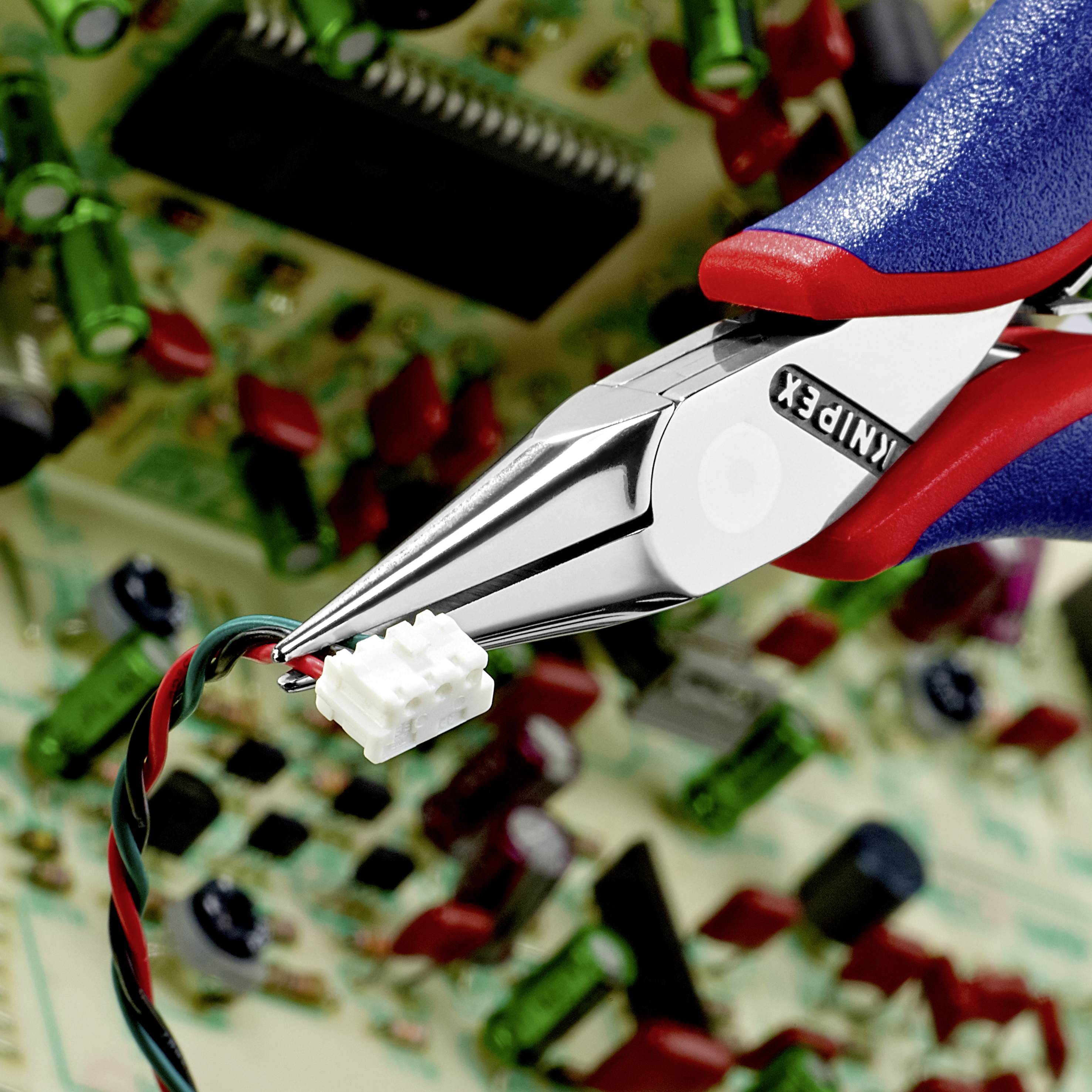 'Nahaufnahme einer Zange mit rot-blauem Griff, die einen kleinen weißen Stecker halt. Im Hintergrund unscharfe elektronische Platine. Hauptaugenmerk auf dem Steckerverbindungsvorgang.'